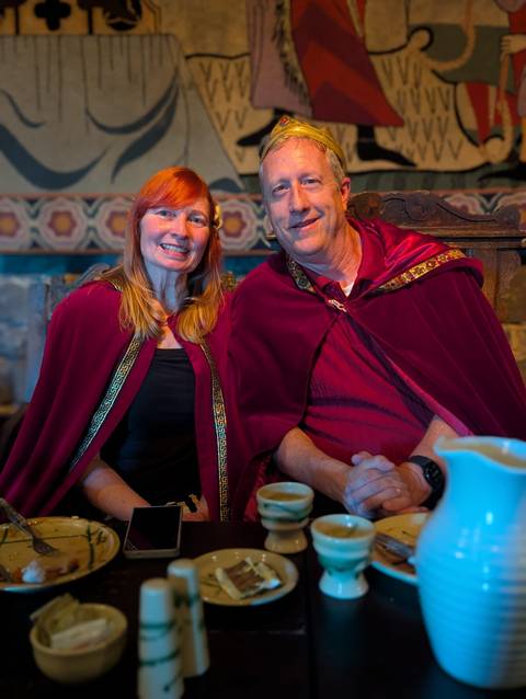       Smiling couple in medieval-style cloaks seated indoors at a rustic banquet setting
  