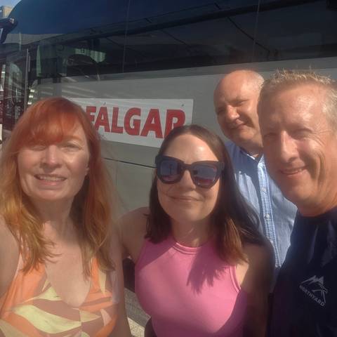       Casual selfie of four travellers smiling in front of a tour coach on a sunny day
  