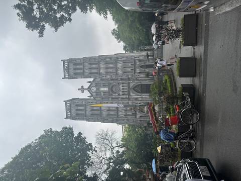       Gothic-style cathedral facade in a city square with rickshaw, pedestrians and umbrellas on a grey day
  