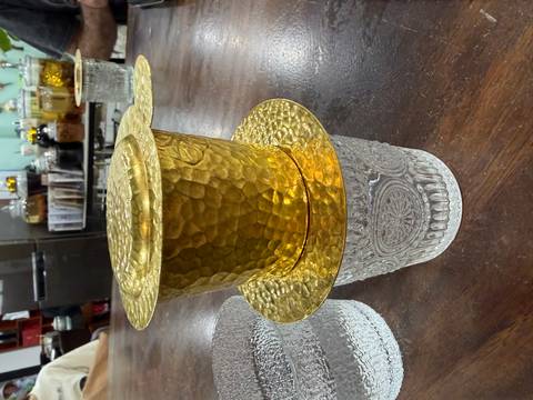       Close shot of a hammered-gold Vietnamese phin coffee filter resting atop an ornate glass tumbler on a wooden table.
  