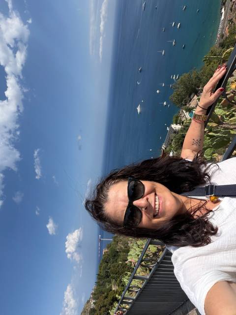       Selfie of a smiling woman wearing sunglasses at a high seaside lookout with deep-blue Mediterranean waters and yachts below
  