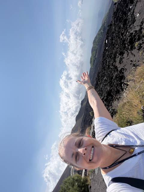       Partial view of a woman’s outstretched arm pointing over dark volcanic rock fields and rolling slopes under a bright sky
  