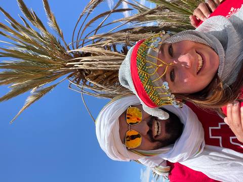       Couple dressed in desert headscarves laughing beneath a tall palm tree and blue sky.
  