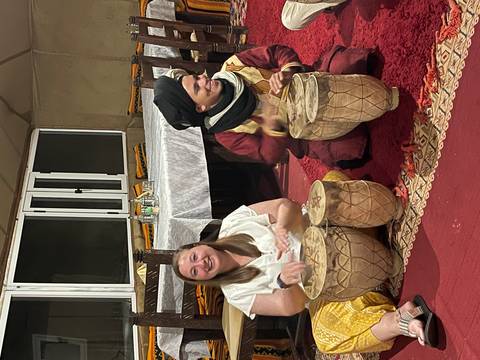       Two people playing hand drums inside a Bedouin-style desert tent at night.
  