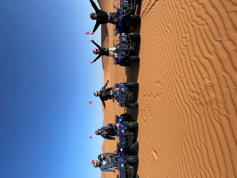       Four riders celebrating on quad bikes atop golden desert dunes under a clear sky.
  