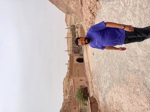       Traveller standing on a dusty track outside an earthen kasbah structure on an overcast desert day.
  