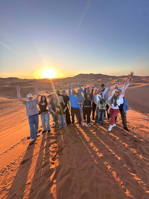       Happy tour group poses on a dune at sunset, arms raised against rippled desert sands.
  