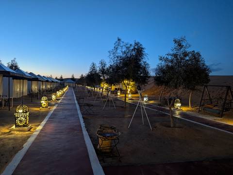       Evening view of a desert camp walkway lined with illuminated Moroccan lanterns and tents beside sand dunes
  