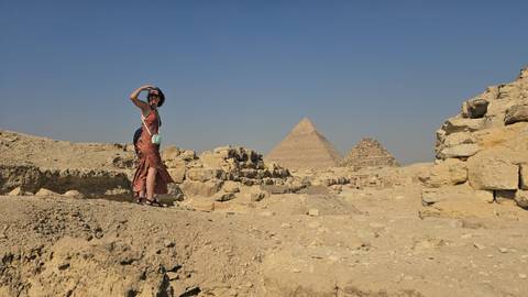       A lone traveller strikes a pose atop sandy ruins with the pyramids rising in the hazy distance.
  