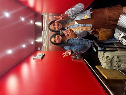       Two friends take a cheerful mirror selfie in a red elevator holding up peace signs with luggage nearby
  