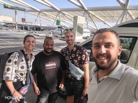       Selfie of three travelers and their driver outside an airport terminal under a modern canopy.
  