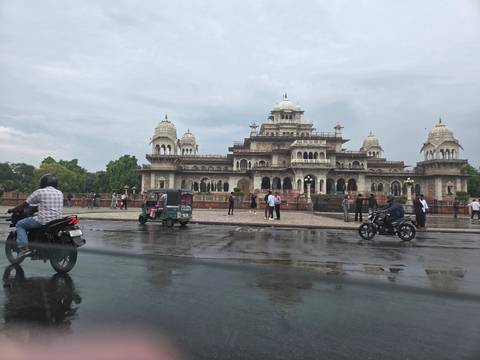       Albert Hall Museum Jaipur viewed across wet street with passing motorbikes and rickshaw.
  