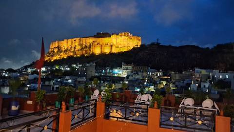       Mehrangarh Fort glows golden above the blue houses of Jodhpur under a deep evening sky.
  