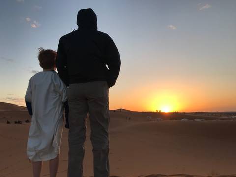       Adult and child stand atop a sand dune watching a brilliant desert sunrise.
  