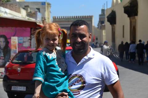       Smiling man carries a little girl in a bustling Moroccan medina street.
  