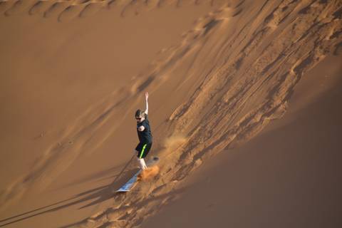       Boy sandboards down a steep dune leaving a trail of orange dust behind him.
  