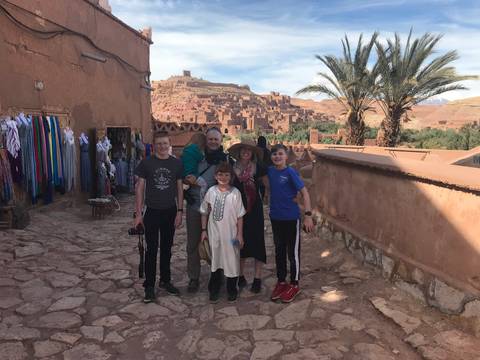       Family stands on a cobbled lane with the clay ksar of Ait Benhaddou and palm trees in the background.
  