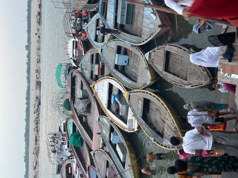       Many wooden rowboats moored along busy riverside ghat with people preparing to board
  