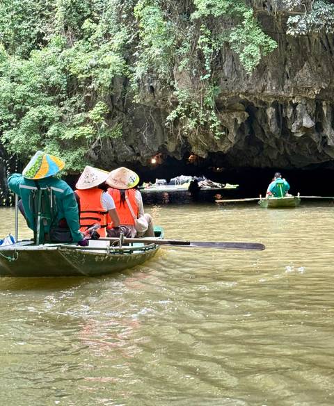       Tourists in conical hats paddle a small boat toward a dark limestone cave entrance on a calm river.
  