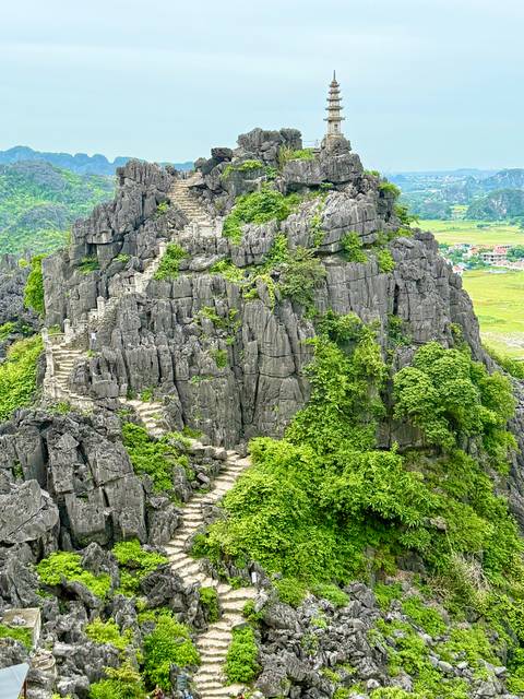       Steep rocky peak covered with greenery and zig-zag stone steps overlooking rice fields below.
  