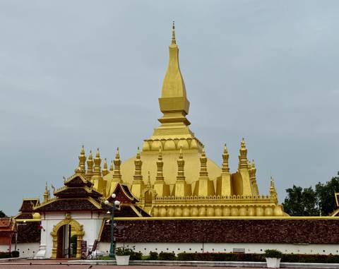       Golden stupa complex rises against a grey sky with ornate roofs and spires.
  