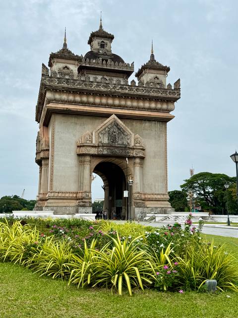       Visitors walk under the ornate arch of Patuxai monument surrounded by gardens.
  