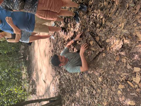       Guide demonstrates a narrow Cu Chi tunnel opening while travelers observe in a wooded area.
  