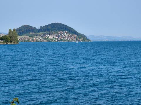       Blue lake with distant hillside village and green headland under hazy sky.
  