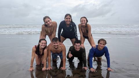       Seven friends build a human pyramid on a wet beach with grey waves rolling behind them.
  