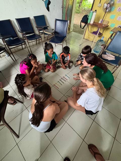       Volunteers and local children sit in a circle on tiled floor playing an educational stick game.
  