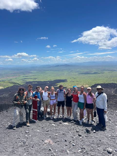       Hikers pose atop a volcanic ridge with vast green plains and black lava fields stretching below.
  