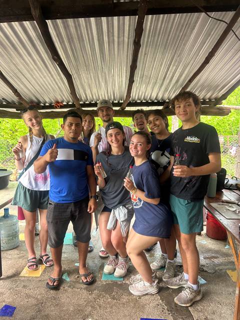      Volunteers hold finished glass bottle crafts while posing in a rustic shelter.
  
