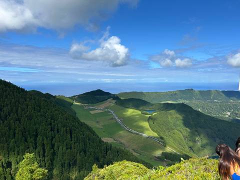       Panoramic viewpoint over rolling green hills, farmland, and a distant blue ocean beneath a bright sky with scattered clouds.
  