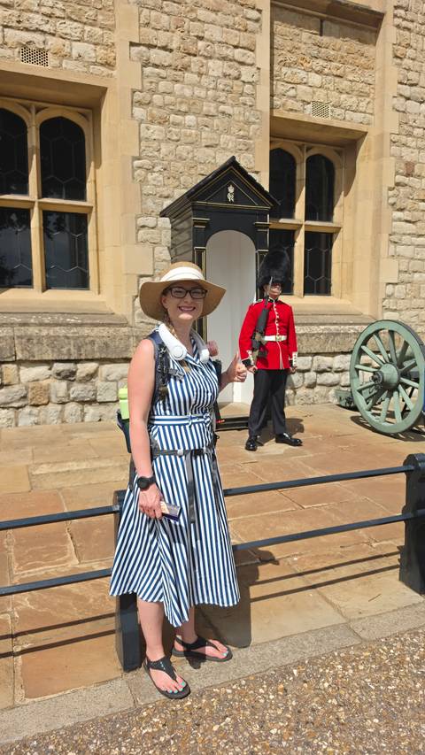       Female tourist giving a thumbs-up beside a stoic British guard in red uniform and a historic cannon outside a stone fortress building
  