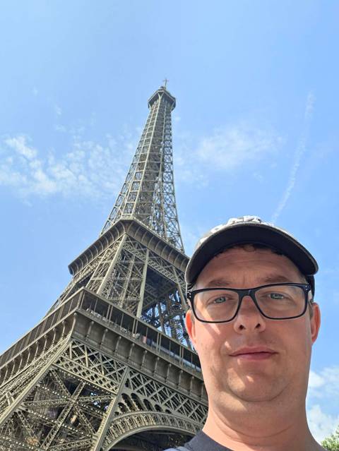       Partial selfie of a man wearing glasses with the Eiffel Tower soaring into a blue sky behind him
  