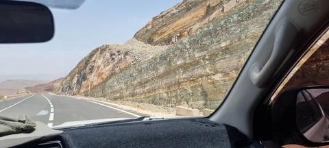       Mountain road seen through a car windshield alongside multicolored rock strata.
  