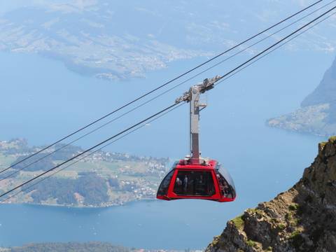       Modern red cable car travels high above a sparkling blue lake and patchwork farmland.
  