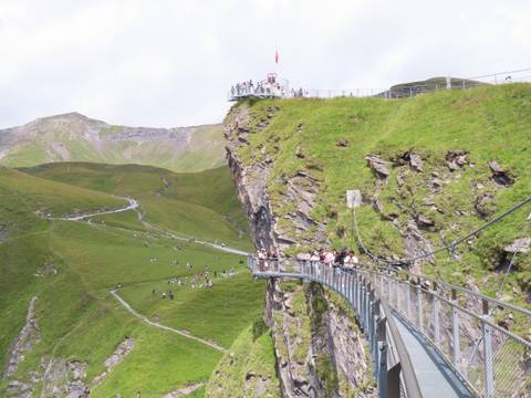       Crowds walk along a dramatic suspended walkway clinging to a grassy cliff face.
  