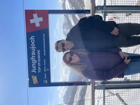       Smiling couple pose beside Jungfraujoch ‘Top of Europe’ sign with snowy alpine peaks behind.
  