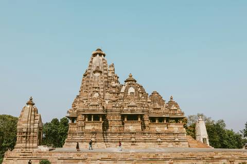       Full view of a soaring Khajuraho temple shikhara set against a clear blue sky with tiny visitors at the base.
  