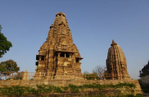       Side profile of two sandstone temples at Khajuraho surrounded by greenery on a sunny afternoon.
  