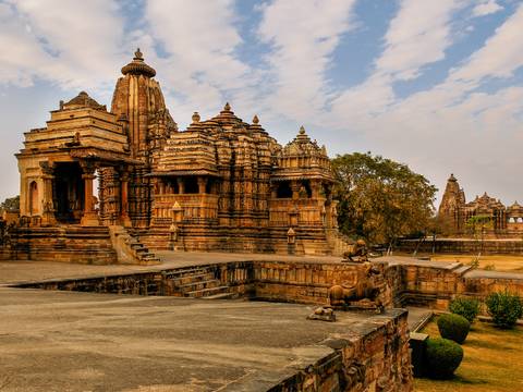       Expansive courtyard of Khajuraho temple complex with ornate sandstone shrines and stone guardian statues under a partly cloudy sky.
  