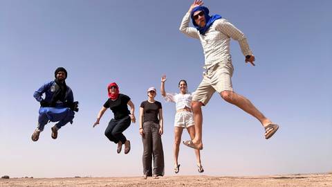       Five travelers in desert scarves leap joyfully against a clear blue sky on sandy terrain.
  