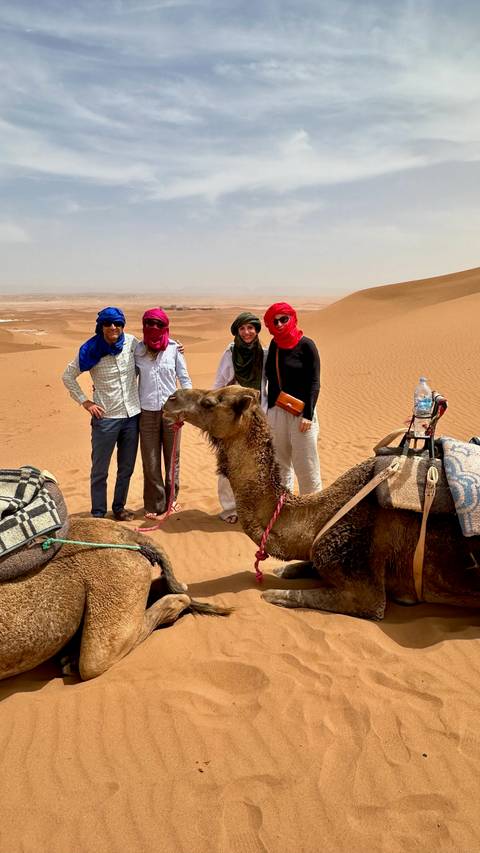       Travelers pose beside a kneeling camel draped in blankets with desert vistas beyond.
  