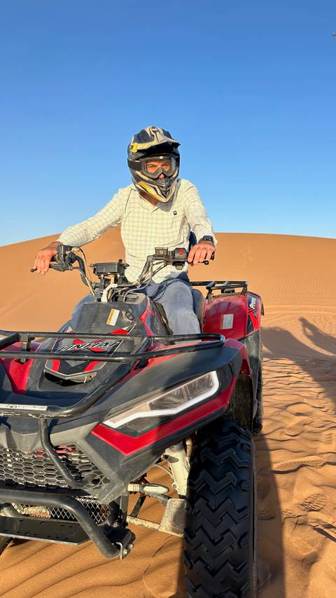       Man drives a red quad bike over smooth orange sand dunes under bright sun.
  