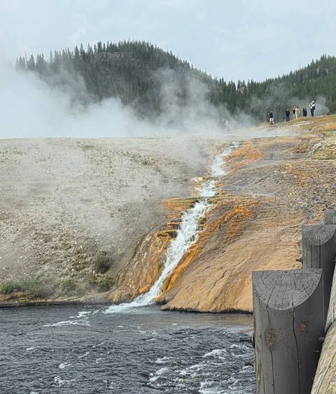       Steaming geothermal run-off flowing over vivid orange mineral deposits in a Yellowstone thermal area with visitors in the distance.
  