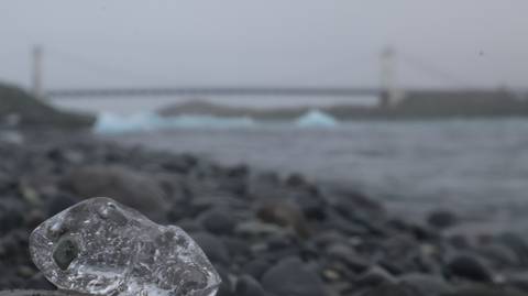       Out-of-focus ice fragment on pebbled shore with bridge and Jokulsarlon lagoon behind in fog.
  