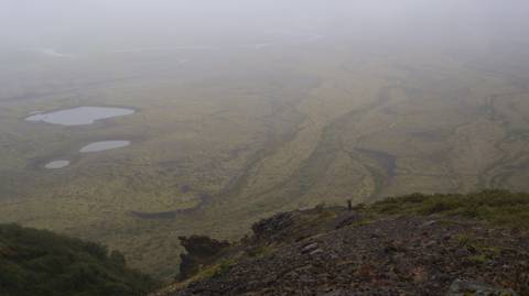       Hiker in misty panoramic view over Icelandic highlands with rivers and ponds far below.
  