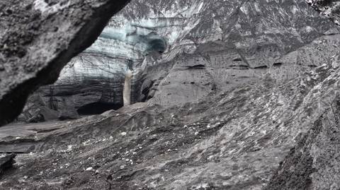       Dark volcanic ash gorge revealing vivid blue ice cave and waterfall of Skaftafell glacier.
  