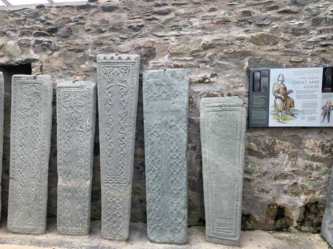       A row of intricately carved grey medieval grave slabs displayed against a rough stone wall with an information panel.
  